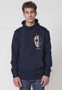 Sudadera azul marino de algodón con un bolsillo frontal, presenta un gráfico de calavera y costuras decorativas, complementada por detalles de cordón.