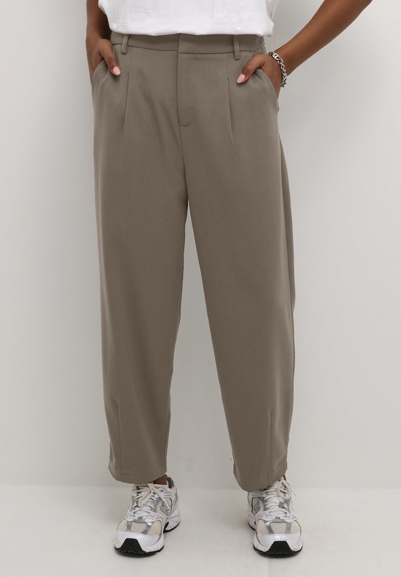 Pantalon beige sur mesure avec plis, doté de poches latérales et d'une coupe décontractée. Porté avec des baskets grises et blanches, sur un fond clair.