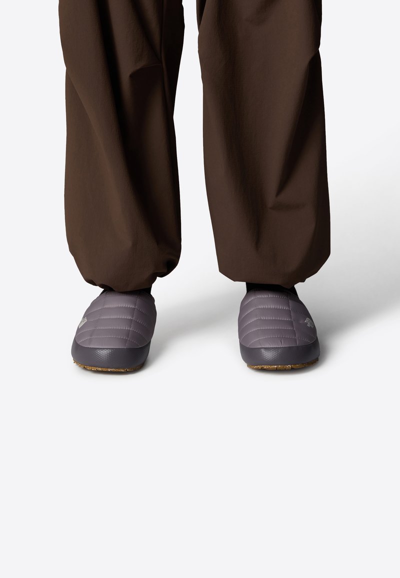 Zapatos de casa morados acolchados con una superficie texturizada, suelas de goma y pantalones marrones oscuros. Los zapatos tienen un logo en el lateral.