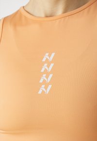 Oranžový sportovní tílko s hladkou texturou a minimem švů, s bílým logem vertikálně umístěným ve středu.