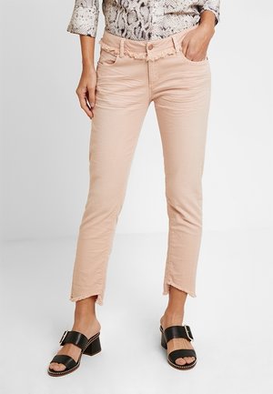 Vrouw in lichtroze jeans met versleten zoom, slangenprint top en zwarte sandalen met hakken en gespen, staand met één hand in haar zak.