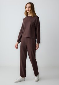 Felpa marrone con dettagli borchiati, abbinata a pantaloni tapered marroni. Sneakers bianche completano l'outfit. Tessuto morbido, design casual.