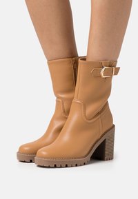 Botas de tobillo de cuero marrón claro con cremallera, tacón en bloque texturizado y detalle de hebilla decorativa. Acabado suave con punta redondeada y suela con relieve.