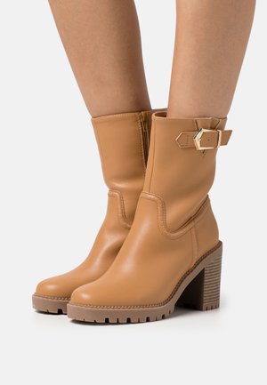 Botas de tobillo de cuero marrón claro con cremallera, tacón en bloque texturizado y detalle de hebilla decorativa. Acabado suave con punta redondeada y suela con relieve.