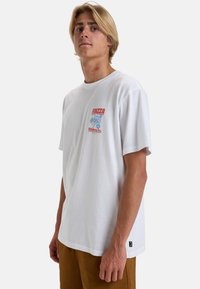Witte katoenen t-shirt met een ronde hals, voorzien van een rood en blauw grafisch patroon op de linkerkant van de borst, en korte mouwen.