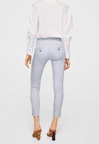 Pruhované kalhoty v bledě modré a bílé barvě, slim fit střih, hladký materiál, zadní kapsy s modrými knoflíky, zkrácená délka, nošeny s hnědými podpatky.