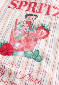 Betty Boop assise sur une boisson rose à la fraise avec de la glace, entourée de fraises perlées et de feuilles de menthe sur un tissu rayé.