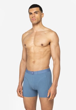 Uomo muscoloso in piedi che indossa boxer blu con logo sulla fascia in vita, rivolto verso l'obiettivo su uno sfondo bianco semplice.