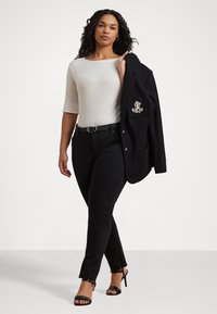 Blazer preto com botões dourados e um logotipo bordado, combinado com uma blusa branca de mangas três quartos e calças justas pretas. Sapatos de salto pretos.