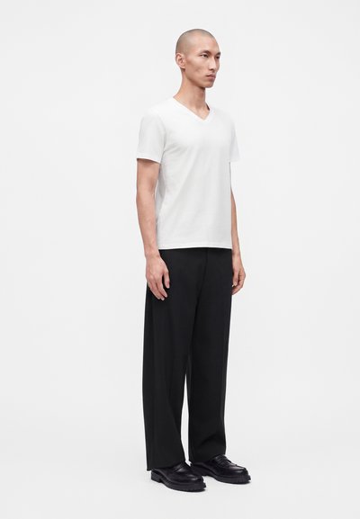 Homme debout de profil portant un t-shirt blanc à manches courtes en V, un pantalon large noir et des chaussures noires sur un fond blanc uni.