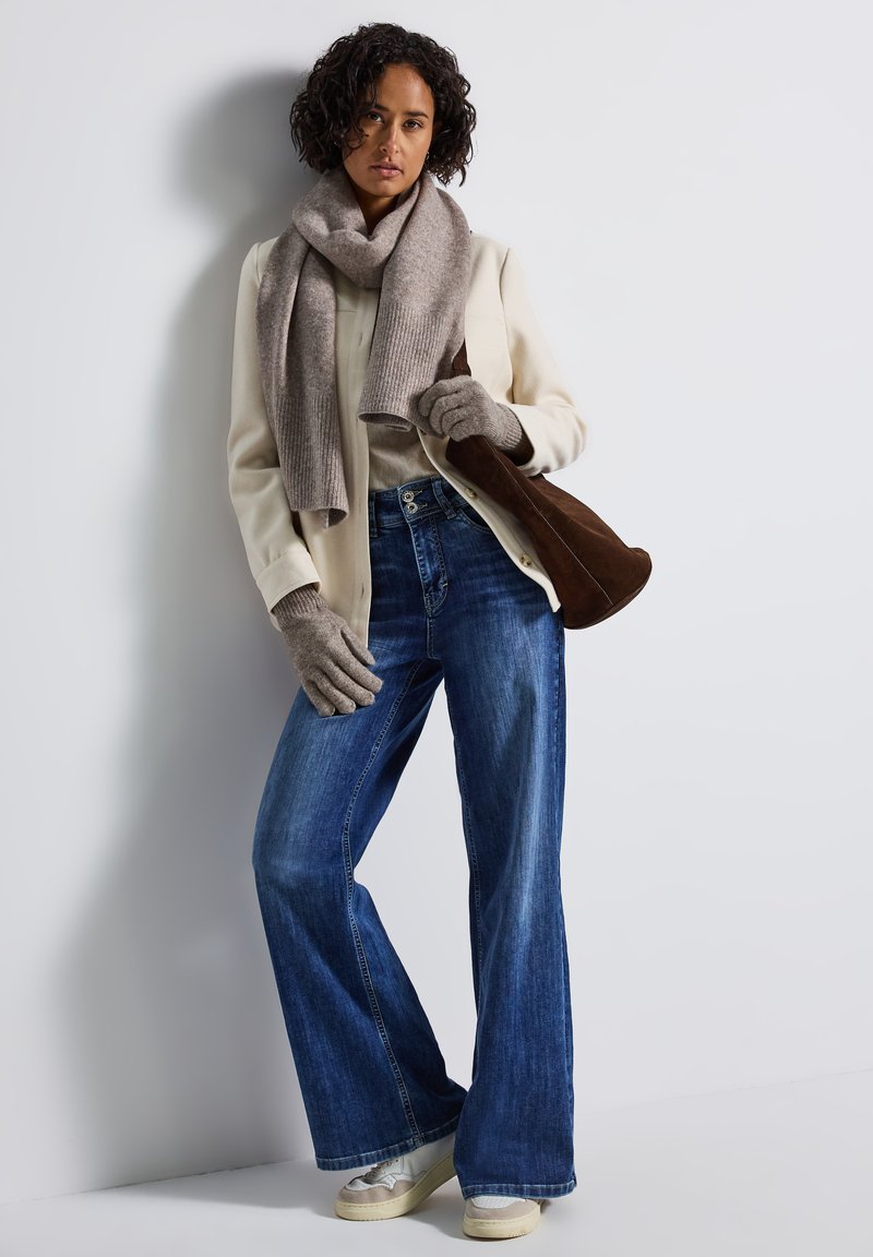 Laivastonsiniset leveät farkut, kerma neule, beige huivi, harmaat hanskat ja ruskea mokkabagsi. Malli seisoo yksinkertaista taustaa vasten.