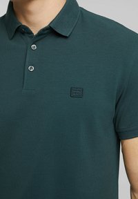Donkergroen poloshirt met een gestructureerde stof, een knoopsluiting met twee knopen en een klein zwart logoplaatje op de borst. Korte mouwen.