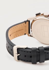 Μαύρο δερμάτινο λουράκι ρολογιού με ασημένια αγκράφα, μερικώς προσαρτημένο σε ασημένια θήκη ρολογιού, που παρουσιάζει ραμμένες άκρες και εσωτερική σήμανση μάρκας.