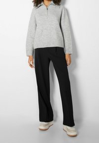 Suéter de punto gris claro con cuello con media cremallera, combinado con pantalones negros de corte holgado y zapatos de plataforma color crema. Texturas suaves y diseño moderno.