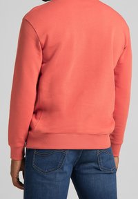 Koraalkleurige sweatshirt met ribgebreide manchetten en onderrand, gemaakt van textuurstof. Draagt met donkerblauwe jeans met een subtiel zakontwerp.