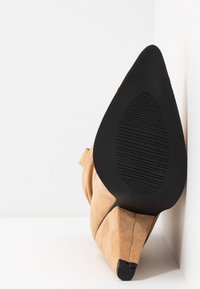 Chaussure à talon haut en daim fauve avec une pointe effilée et une semelle en caoutchouc noire texturée, présentant une silhouette lisse et un léger détail de pli.