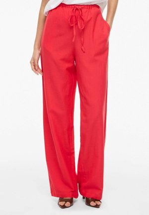 Kvinde iført lyse røde, løstsiddende bukser med elastisk snøre i taljen og sidelommer, kombineret med brune åbne tå-hæle.
