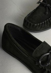 Paire de chaussons mocassins en daim noir avec détails cousus et nœuds en lacets en cuir sur une surface grise.