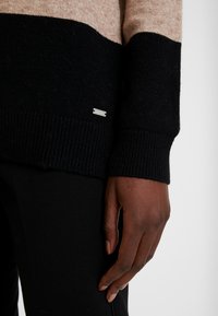 Maglione color block nero e beige con polsini a coste, realizzato in materiale morbido. Presenta un piccolo dettaglio con etichetta argentata sul lato.