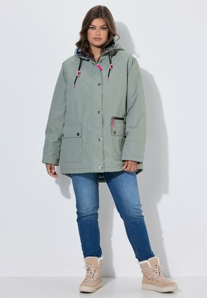 Frau trägt eine hellgrüne Winterjacke mit Kapuze und rosa Akzenten, blaue Jeans und beige Schnürstiefel mit Fellfutter, steht vor weißem Hintergrund.
