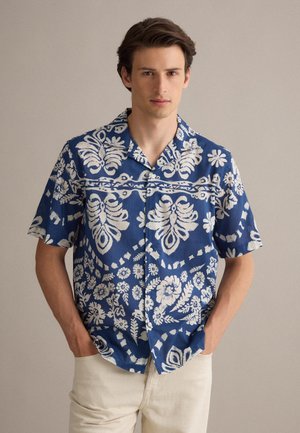 Giovane uomo con camicia blu a maniche corte con motivi floreali bianchi e pantaloni beige, in piedi con le mani nelle tasche davanti a uno sfondo neutro.
