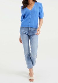 Modrý žebrovaný cardigan s výstřihem do V, krátkými rukávy a zapínáním na knoflíky, doplněný světle modrými přiléhavými džíny a neutrálními sandály.