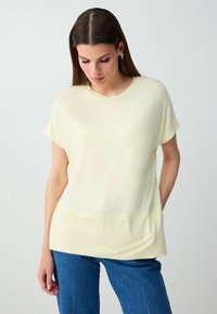 Femme aux cheveux bruns et raides portant un haut jaune pâle à manches courtes et un jean bleu, regardant vers le bas avec une main derrière le dos.