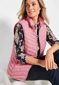 Helle rosa gesteppte Weste mit hohem Kragen und einem Reißverschluss vorne. Darunter ein schwarzes Hemd mit floralen Mustern, kombiniert mit dunklen Hosen.