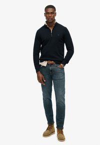 Marineblauwe pullover met een half-rits kraag en logo, gecombineerd met lichtblauwe jeans en tan kleurige veterschoenen. Casual pasvorm met een gelaagde look.