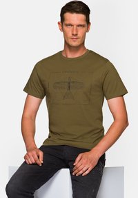 Oliwkowa bawełniana koszulka z krótkim rękawem, z czarnym nadrukiem schematu samolotu z przodu. Stylizowana z ciemnymi dżinsami.