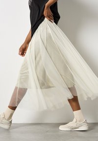 Durchsichtiges, irisierendes Tüllrock in hellem Creme mit Falten-Design. Kombiniert mit einem schwarzen Shirt, beigen Knöchelsocken und weißen Turnschuhen.