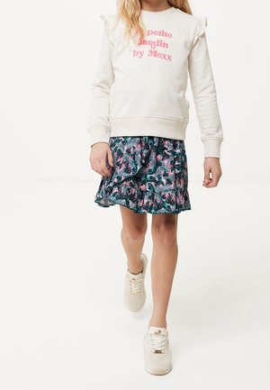 Fille portant un sweat-shirt blanc avec un texte rose et une jupe à imprimé tropical bleu marine, marchant en avant en baskets blanches.