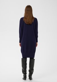 Langärmliges, dunkel marineblaues Pulloverkleid mit geripptem Saum und Seitentaschen, kombiniert mit kniehohen schwarzen Lederstiefeln. Glatte Textur.