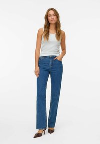 Woman standing with hand in pocket wearing white sleeveless top, blue straight-leg jeans, and brown pointed-toe heels with studs.