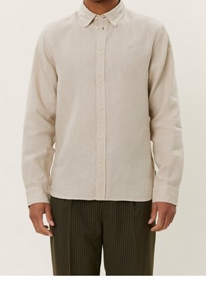 Homme portant une chemise beige à manches longues avec col et boutons et un pantalon foncé à fines rayures, debout devant un fond uni.