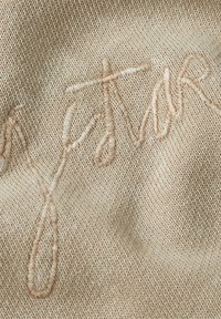 Beige textured fabric featuring raised, embroidered script in a lighter beige, creating a subtle tonal contrast against the background.