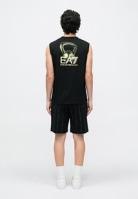 Must musta varrukateta särk kuldse logoga seljal, kombineeritud mustade lühikeste pükstega, millel on korduv logomuster; kantud valgete sokkide ja tossudega.