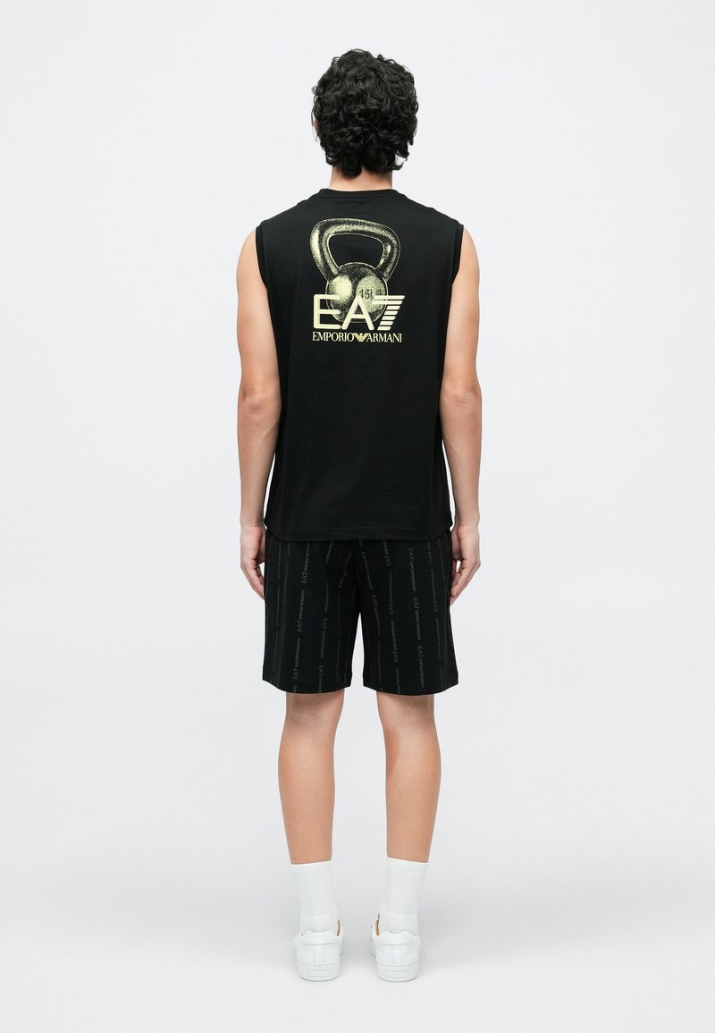 Must musta varrukateta särk kuldse logoga seljal, kombineeritud mustade lühikeste pükstega, millel on korduv logomuster; kantud valgete sokkide ja tossudega.