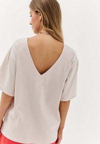 Světlé béžové tričko s hlubokým výstřihem na zádech, krátkými rukávy a jemnou, měkkou texturou. Jednoduchý a uvolněný design.