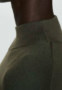 Personne à la peau foncée portant un pull en maille vert olive à col montant, mettant en avant la zone du cou et des épaules sur un fond uni.