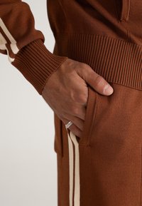 Mână în buzunarul de la pantaloni maro, purtând un pulover maro asortat cu dungi crem pe mânecă și un inel de argint pe degetul inelar.