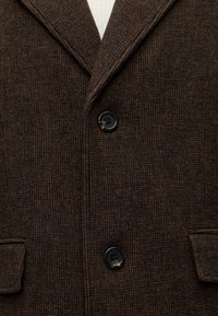 Gros plan d'un manteau en laine marron à carreaux avec deux boutons noirs et des poches à rabat, porté sur un pull crème.