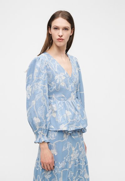 Femme aux longs cheveux bruns portant un haut à manches longues bleu et une jupe avec des motifs blancs de vie marine, debout devant un fond blanc uni.