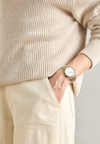Beżowy sweter z prążkowanego dzianiny, w połączeniu z jasnobeżowymi spodniami z boczną kieszenią. Złoty siatkowy zegarek z białą tarczą na nadgarstku.