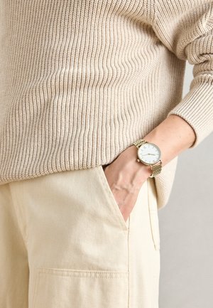 Beige ribbattu neule, yhdistettynä vaaleanbeigeihin housuihin, joissa on sivutasku. Kultainen verkkokello valkoisella kellotaululla ranteessa.