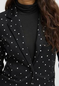 Svart kavaj med små vita hjärtan som bärs över en svart turtleneck, som visar övre delen av överkroppen och vågigt brunt hår.