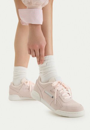 Osoba poprawiająca białe prążkowane skarpety, nosząca jasnoróżowe zamszowe trampki ze sznurowadłami na białym tle.