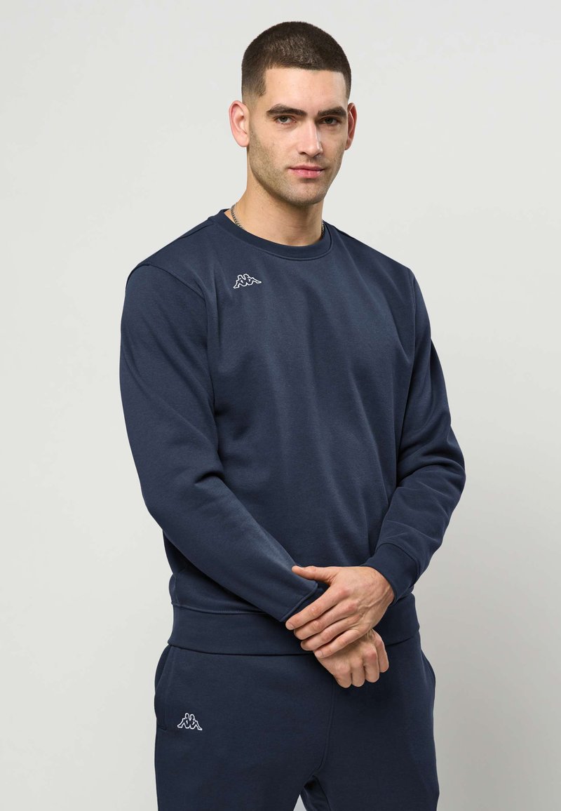 Homme aux cheveux courts portant un sweat-shirt bleu marine et un pantalon assorti, debout devant un fond uni clair, les mains jointes.
