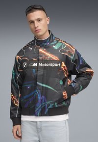 Puma BMW Motorsport-jacka som har en svart tyg med flerfärgade elektriska mönster och vita detaljer; innehåller logotyp och uppstående krage.