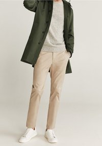 Vihreä takki, harmaa neule, beiget housut ja valkoiset lenkkarit. Takissa on napit ja huppu; neuleessa on tekstuuri. Housut ovat kapealahkeiset.